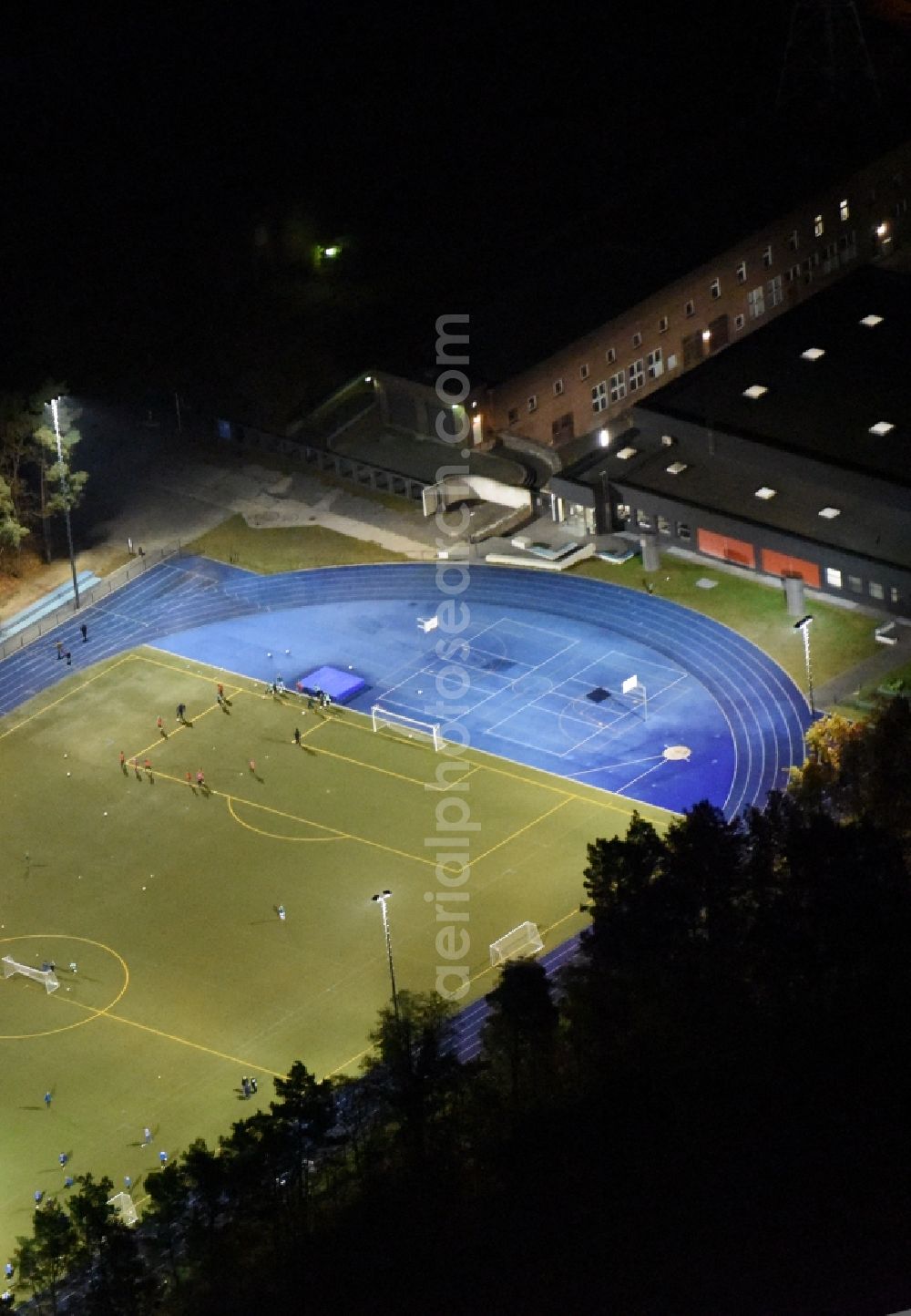 Kleinmachnow at night from above - Night image of the sports facilities of the Berlin Brandenburg International School and residential buildings in Kleinmachnow in the state of Brandenburg. The BBIS includes a football pitch and athletics facilities in a distinct blue colour. Residential buildings and estates as well as a primary school are surrounded by trees and woods