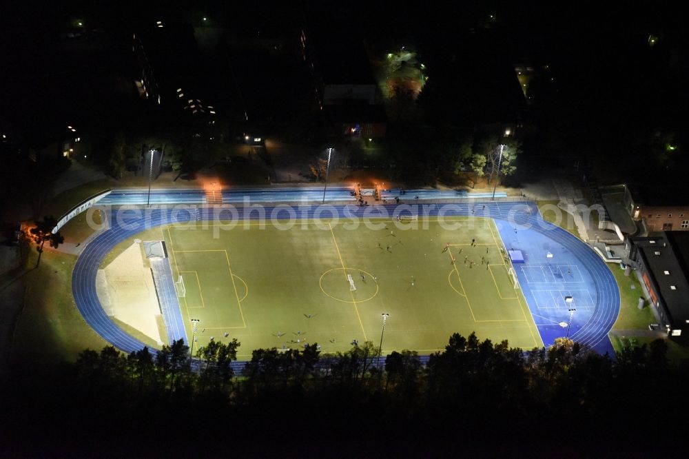 Kleinmachnow at night from the bird perspective: Night image of the sports facilities of the Berlin Brandenburg International School and residential buildings in Kleinmachnow in the state of Brandenburg. The BBIS includes a football pitch and athletics facilities in a distinct blue colour. Residential buildings and estates as well as a primary school are surrounded by trees and woods