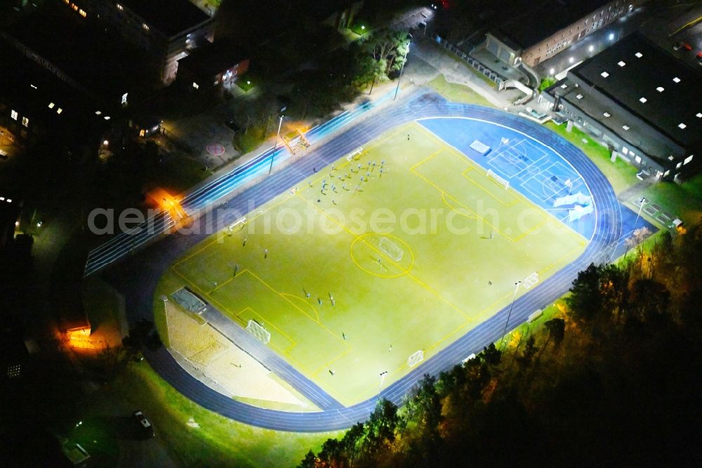 Aerial image at night Kleinmachnow - Night lighting Sports facilities of the Berlin Brandenburg International School and residential buildings in Kleinmachnow in the state of Brandenburg. The BBIS includes a football pitch and athletics facilities in a distinct blue colour. Residential buildings and estates as well as a primary school are surrounded by trees and woods