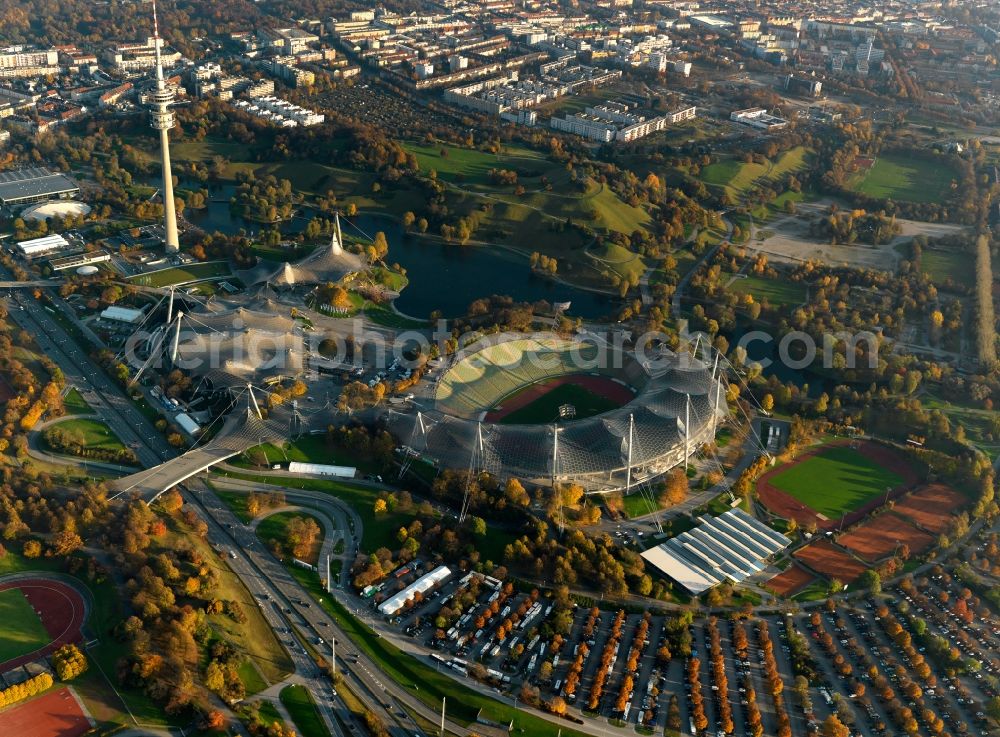 München from above - The Olympic Park in Munich in the state of Bavaria. In the foreground lies the Olympic Stadium that is today used for cultural events and concerts. Behind it, the TV tower, the Olympic Tower, one of the landmarks of the city is visible. The large park and lake form one of the largest open spaces in Munich