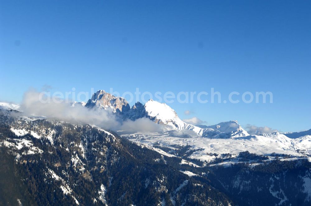 Aerial photograph SEIS - Winterlich verschneite Seiser Alm mit dem Schlern Gebirgsmassiv der Dolomiten. Der Schlern (2.563 m, ital. Sciliar) ist ein Berg in den Dolomiten/Alpen in Südtirol, Italien. Trotz seiner geringen Höhe gilt er u.a. auf Grund seiner charakteristischen Form mit den beiden vorgelagerten Bergspitzen, der Santner- (2.413 m s.l.m.) und der Euringerspitze (2.394 m s.l.m.), als Wahrzeichen Südtirols. An den Schlern angelehnt ist die Seiser Alm, die höchstgelegene Hochweide Europas. 1974 wurde das umgebende Gebiet zum Naturpark erklärt, heute Teil des Naturparks Schlern-Rosengarten.Das Schlern-Massiv entstand wie die übrigen Berge der Dolomiten gegen Ende des Erdaltertums, in der sog. Permzeit. In der Tethys, die damals diese Landmassen bedeckte, sanken während einer langen Zeitspanne Ablagerungen von kleinen kalkhaltigen Tieren auf den Grund. Temperatur- und Lichtverhältnisse waren wahrscheinlich die Ursache, dass sich Korallenriffe ansiedelten. So bauten sich im Lauf von Jahrmillionen riesige Korallenbauten von tausenden Metern auf. Druck verdichtete langsam das Kalkgebilde. Durch Absinken der darunter liegenden Bozner Quarzporphyrplatte bildete sich die typische Form des Schlerns. Dann, zu Beginn des Erdmittelalters, erhob sich das Gebiet aus der Tethys. Die Afrikanische Platte schob sich langsam unter die Eurasische Platte und die Alpen falteten sich auf. Durch Erosion erlangte der Schlern schließlich sein heutiges Gesicht.