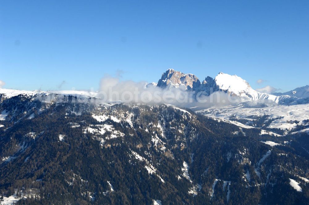SEIS from above - Winterlich verschneite Seiser Alm mit dem Schlern Gebirgsmassiv der Dolomiten. Der Schlern (2.563 m, ital. Sciliar) ist ein Berg in den Dolomiten/Alpen in Südtirol, Italien. Trotz seiner geringen Höhe gilt er u.a. auf Grund seiner charakteristischen Form mit den beiden vorgelagerten Bergspitzen, der Santner- (2.413 m s.l.m.) und der Euringerspitze (2.394 m s.l.m.), als Wahrzeichen Südtirols. An den Schlern angelehnt ist die Seiser Alm, die höchstgelegene Hochweide Europas. 1974 wurde das umgebende Gebiet zum Naturpark erklärt, heute Teil des Naturparks Schlern-Rosengarten.Das Schlern-Massiv entstand wie die übrigen Berge der Dolomiten gegen Ende des Erdaltertums, in der sog. Permzeit. In der Tethys, die damals diese Landmassen bedeckte, sanken während einer langen Zeitspanne Ablagerungen von kleinen kalkhaltigen Tieren auf den Grund. Temperatur- und Lichtverhältnisse waren wahrscheinlich die Ursache, dass sich Korallenriffe ansiedelten. So bauten sich im Lauf von Jahrmillionen riesige Korallenbauten von tausenden Metern auf. Druck verdichtete langsam das Kalkgebilde. Durch Absinken der darunter liegenden Bozner Quarzporphyrplatte bildete sich die typische Form des Schlerns. Dann, zu Beginn des Erdmittelalters, erhob sich das Gebiet aus der Tethys. Die Afrikanische Platte schob sich langsam unter die Eurasische Platte und die Alpen falteten sich auf. Durch Erosion erlangte der Schlern schließlich sein heutiges Gesicht.