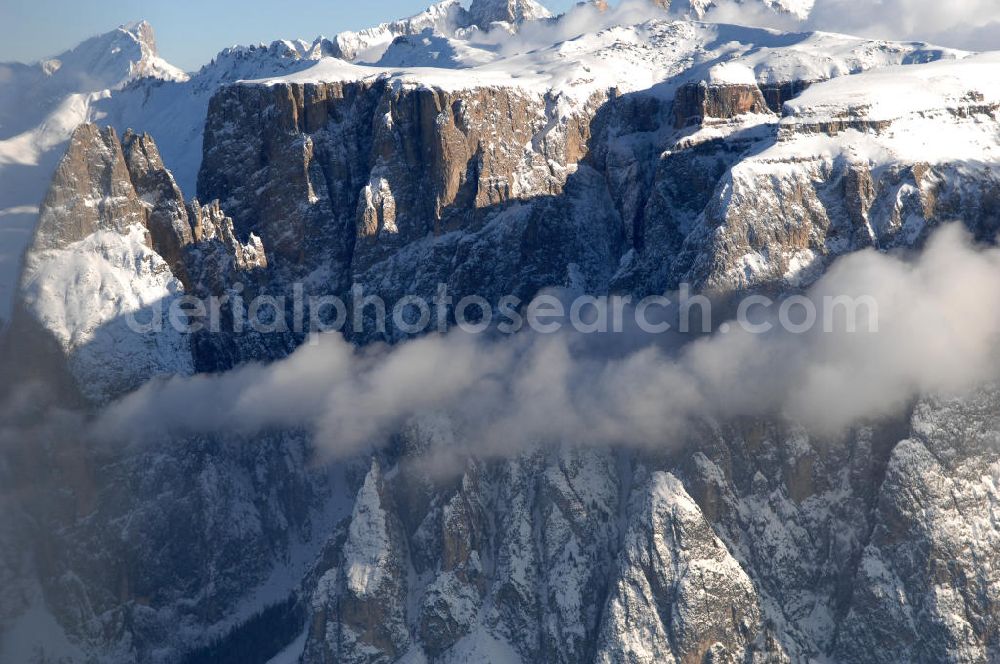 SEIS from the bird's eye view: Winterlich verschneite Seiser Alm mit dem Schlern Gebirgsmassiv der Dolomiten. Der Schlern (2.563 m, ital. Sciliar) ist ein Berg in den Dolomiten/Alpen in Südtirol, Italien. Trotz seiner geringen Höhe gilt er u.a. auf Grund seiner charakteristischen Form mit den beiden vorgelagerten Bergspitzen, der Santner- (2.413 m s.l.m.) und der Euringerspitze (2.394 m s.l.m.), als Wahrzeichen Südtirols. An den Schlern angelehnt ist die Seiser Alm, die höchstgelegene Hochweide Europas. 1974 wurde das umgebende Gebiet zum Naturpark erklärt, heute Teil des Naturparks Schlern-Rosengarten.Das Schlern-Massiv entstand wie die übrigen Berge der Dolomiten gegen Ende des Erdaltertums, in der sog. Permzeit. In der Tethys, die damals diese Landmassen bedeckte, sanken während einer langen Zeitspanne Ablagerungen von kleinen kalkhaltigen Tieren auf den Grund. Temperatur- und Lichtverhältnisse waren wahrscheinlich die Ursache, dass sich Korallenriffe ansiedelten. So bauten sich im Lauf von Jahrmillionen riesige Korallenbauten von tausenden Metern auf. Druck verdichtete langsam das Kalkgebilde. Durch Absinken der darunter liegenden Bozner Quarzporphyrplatte bildete sich die typische Form des Schlerns. Dann, zu Beginn des Erdmittelalters, erhob sich das Gebiet aus der Tethys. Die Afrikanische Platte schob sich langsam unter die Eurasische Platte und die Alpen falteten sich auf. Durch Erosion erlangte der Schlern schließlich sein heutiges Gesicht.