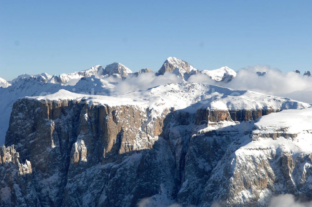Aerial image SEIS - Winterlich verschneite Seiser Alm mit dem Schlern Gebirgsmassiv der Dolomiten. Der Schlern (2.563 m, ital. Sciliar) ist ein Berg in den Dolomiten/Alpen in Südtirol, Italien. Trotz seiner geringen Höhe gilt er u.a. auf Grund seiner charakteristischen Form mit den beiden vorgelagerten Bergspitzen, der Santner- (2.413 m s.l.m.) und der Euringerspitze (2.394 m s.l.m.), als Wahrzeichen Südtirols. An den Schlern angelehnt ist die Seiser Alm, die höchstgelegene Hochweide Europas. 1974 wurde das umgebende Gebiet zum Naturpark erklärt, heute Teil des Naturparks Schlern-Rosengarten.Das Schlern-Massiv entstand wie die übrigen Berge der Dolomiten gegen Ende des Erdaltertums, in der sog. Permzeit. In der Tethys, die damals diese Landmassen bedeckte, sanken während einer langen Zeitspanne Ablagerungen von kleinen kalkhaltigen Tieren auf den Grund. Temperatur- und Lichtverhältnisse waren wahrscheinlich die Ursache, dass sich Korallenriffe ansiedelten. So bauten sich im Lauf von Jahrmillionen riesige Korallenbauten von tausenden Metern auf. Druck verdichtete langsam das Kalkgebilde. Durch Absinken der darunter liegenden Bozner Quarzporphyrplatte bildete sich die typische Form des Schlerns. Dann, zu Beginn des Erdmittelalters, erhob sich das Gebiet aus der Tethys. Die Afrikanische Platte schob sich langsam unter die Eurasische Platte und die Alpen falteten sich auf. Durch Erosion erlangte der Schlern schließlich sein heutiges Gesicht.