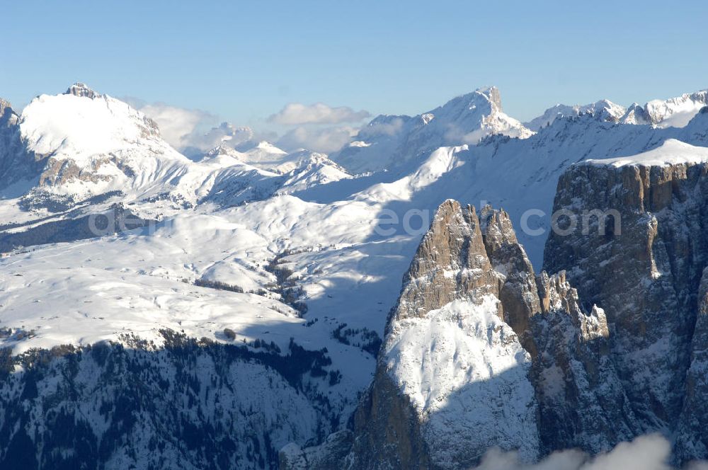 Aerial image SEIS - Winterlich verschneite Seiser Alm mit dem Schlern Gebirgsmassiv der Dolomiten. Der Schlern (2.563 m, ital. Sciliar) ist ein Berg in den Dolomiten/Alpen in Südtirol, Italien. Trotz seiner geringen Höhe gilt er u.a. auf Grund seiner charakteristischen Form mit den beiden vorgelagerten Bergspitzen, der Santner- (2.413 m s.l.m.) und der Euringerspitze (2.394 m s.l.m.), als Wahrzeichen Südtirols. An den Schlern angelehnt ist die Seiser Alm, die höchstgelegene Hochweide Europas. 1974 wurde das umgebende Gebiet zum Naturpark erklärt, heute Teil des Naturparks Schlern-Rosengarten.Das Schlern-Massiv entstand wie die übrigen Berge der Dolomiten gegen Ende des Erdaltertums, in der sog. Permzeit. In der Tethys, die damals diese Landmassen bedeckte, sanken während einer langen Zeitspanne Ablagerungen von kleinen kalkhaltigen Tieren auf den Grund. Temperatur- und Lichtverhältnisse waren wahrscheinlich die Ursache, dass sich Korallenriffe ansiedelten. So bauten sich im Lauf von Jahrmillionen riesige Korallenbauten von tausenden Metern auf. Druck verdichtete langsam das Kalkgebilde. Durch Absinken der darunter liegenden Bozner Quarzporphyrplatte bildete sich die typische Form des Schlerns. Dann, zu Beginn des Erdmittelalters, erhob sich das Gebiet aus der Tethys. Die Afrikanische Platte schob sich langsam unter die Eurasische Platte und die Alpen falteten sich auf. Durch Erosion erlangte der Schlern schließlich sein heutiges Gesicht.