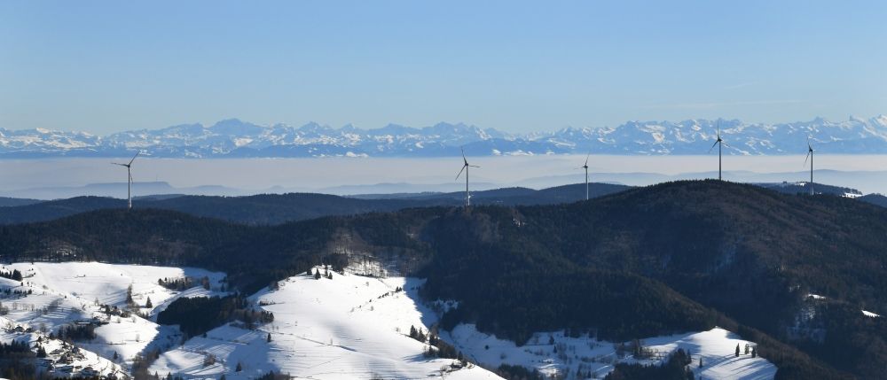 Schopfheim from above - Wintry snowy landscape at the snow covered Rohrenkopf, the local mountain of Gersbach, a district of Schopfheim in Baden-Wuerttemberg. Wind turbines produce renewable energy in the wind farm in the Black Forest. Looking over the mountains to the Swiss Alps