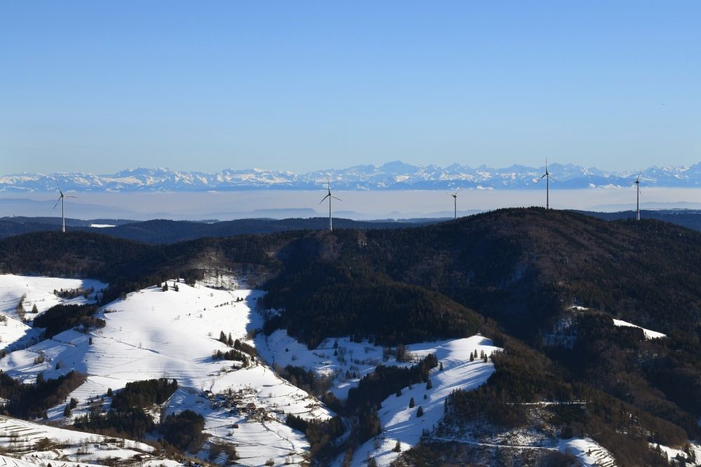 Schopfheim from the bird's eye view: Wintry snowy landscape at the snow covered Rohrenkopf, the local mountain of Gersbach, a district of Schopfheim in Baden-Wuerttemberg. Wind turbines produce renewable energy in the wind farm in the Black Forest. Looking over the mountains to the Swiss Alps